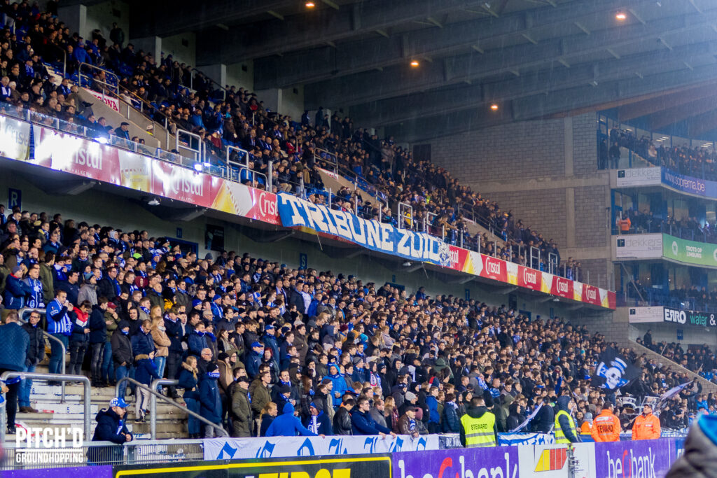 Crowd of KRC Genk supporters in the stands at Cegeka Arena, with fans dressed in blue, banners displayed, and a "Tribune Zuid" sign visible, creating a lively atmosphere.