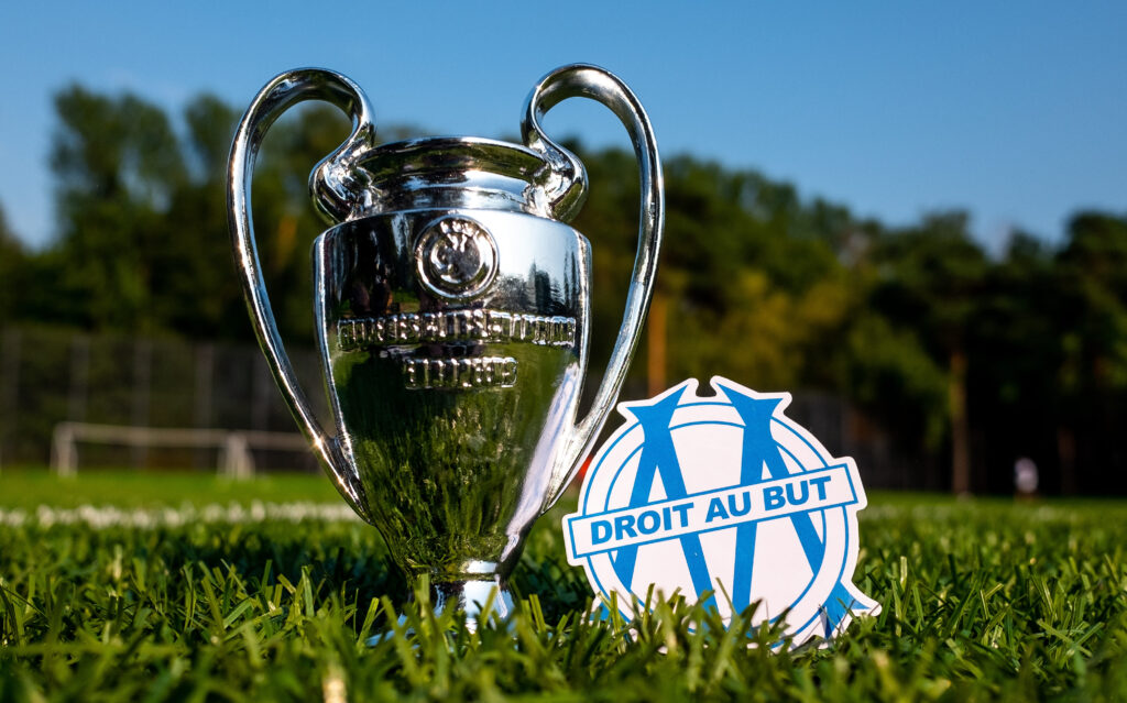 Champions League trophy replica on grass beside the Olympique de Marseille logo, with a blurred football field in the background.