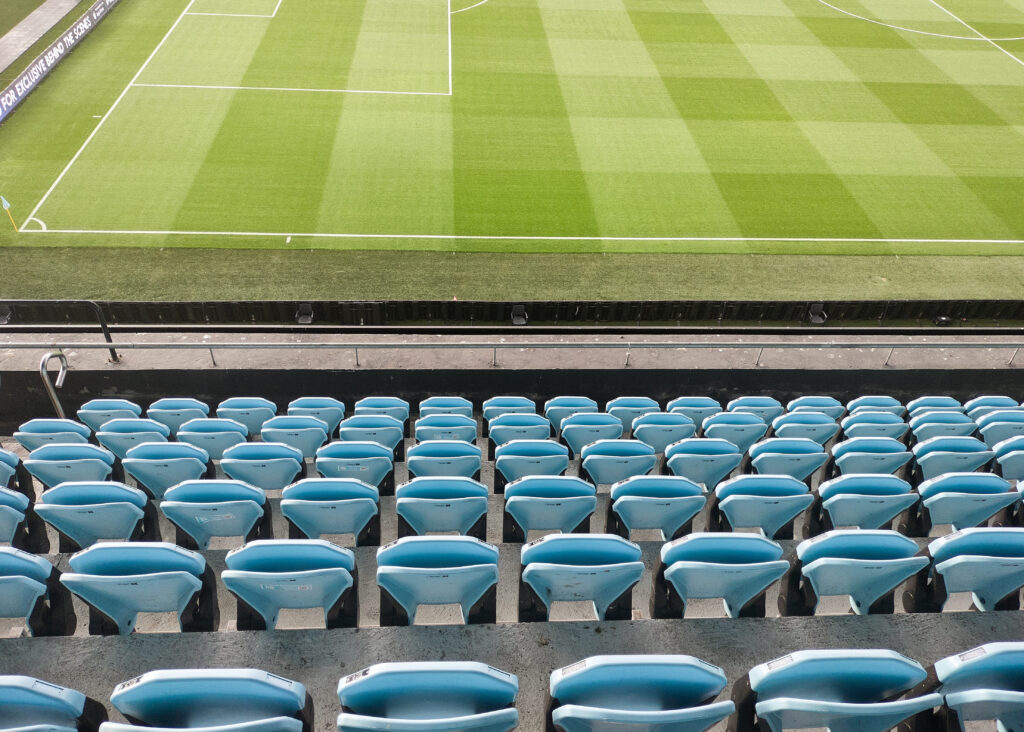 Empty blue stadium seats overlooking a well-maintained football pitch with a checkered grass pattern.